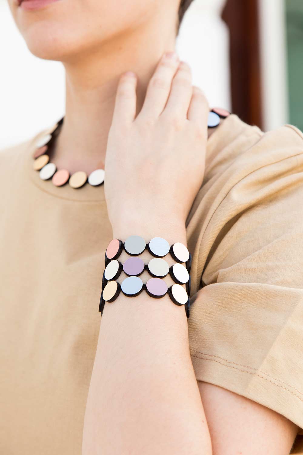 A person wearing a beige shirt is shown from the neck to mid-arm, with their hand resting on their shoulder. They are adorned with elegant jewellery: a beaded necklace and the Abstraction 3 Strand Bracelet by Iskin Sisters, both made of round, multicolored beads in light blue, pink, and beige shades.