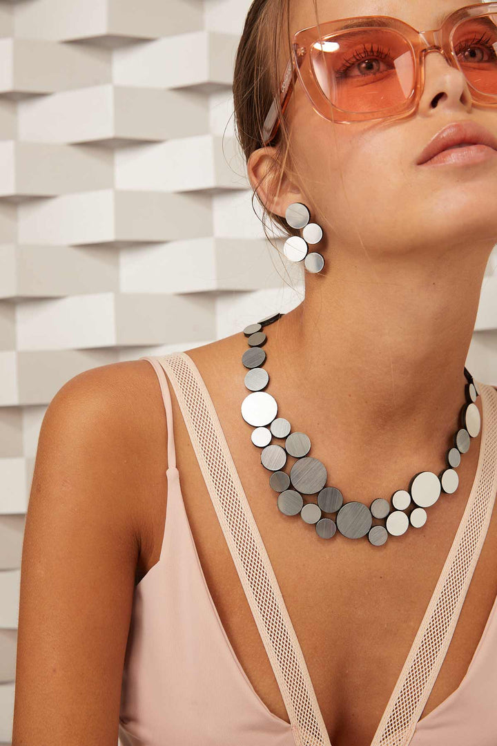 A woman wearing oversized orange sunglasses, a pink sleeveless top with mesh details, and a statement necklace with large circular beads in shades of silver and grey. She also accessorizes her look with the Iskin Sisters' Abstraction Silver Earrings. The background features a textured, geometric design.