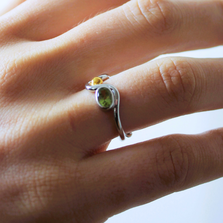 A hand wears the Gallardo & Blaine Designs Adjustable Heartbeat Ring—sterling silver with a larger green and smaller yellow gemstone set on adjacent fingers—against a light background.