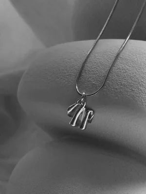 The Little Joy Necklace by Inner Island features a silver pendant with an abstract, wavy design, displayed on a smooth surface in a black and white photo—an elegant piece ideal for everyday wear.