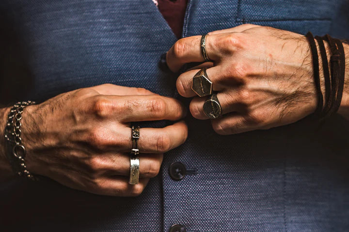 Close-up of a person’s hands adorned with multiple rings and bracelets, buttoning a blue suit jacket. The hands feature visible veins and a mix of handmade jewellery and leather accessories, including the Septentrion Thin Ribbon Ring, adding a fashion-forward and edgy touch to the attire.