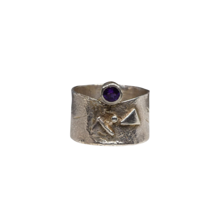 A wide sterling silver ring with a textured surface and an abstract design featuring an amethyst gemstone set into a small circular setting atop the band. The Amethyst Guardian Ring by Caroline Stokesberry-lee includes raised triangular and round shapes, adding to its unique charm.