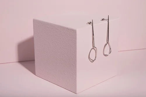 The Ammil Long Earrings by Inner Island, minimalist silver statement pieces, hang on a white textured cube against a pale pink background—a striking addition to any jewellery collection.
