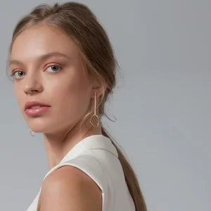 A young woman with light skin and long brown hair tied back looks at the camera, wearing the Ammil Long Earrings by Inner Island with a sleeveless white top against a plain light gray background.