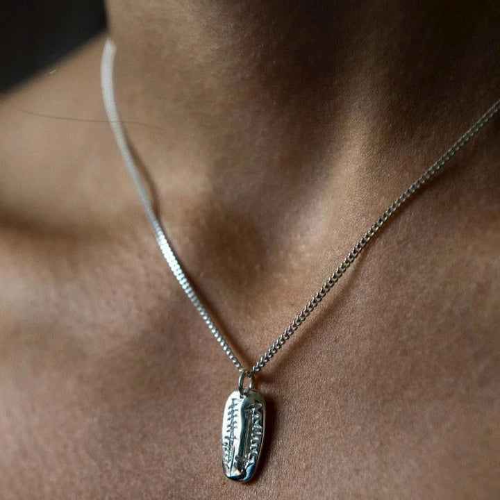 A close-up image of a person wearing a sterling silver pendant necklace with a small, rectangular charm. The Soul Friend (Anam Cara) Carved Pendant by Ogham Treasure features intricate designs with a pattern resembling leaves or feathers, symbolizing an Anam Cara or soul friend. The focus is on the necklace, with the person's neck and upper chest faintly visible.