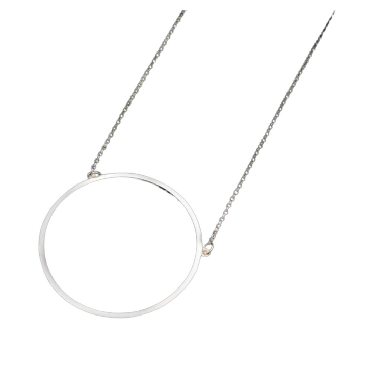 A delicate silver necklace, the Ancaire Silver Neckpiece by Lynsey de Burca, features a simple, hand-forged large circle neckpiece hanging in the center of a fine chain. The minimalist design is elegant and modern, with the sterling silver circle taking up a notable portion of the necklace. The background is plain white.