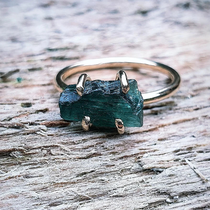 The Raw Green Apatite Gold Filled Ring by Caroline Stokesberry-lee features a prong-set raw green apatite gemstone, beautifully displayed on a weathered wooden surface.