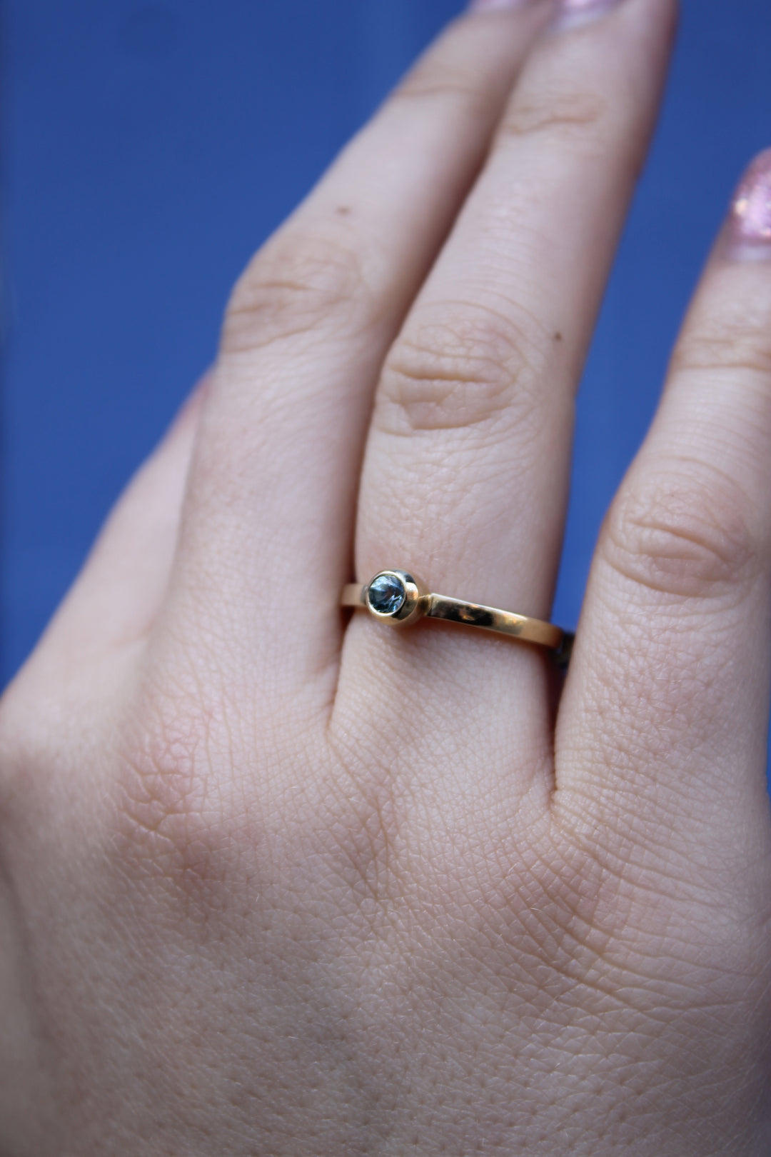 A hand with painted nails wears the Madeleine Azure Drop 9ct Yellow Gold Ring, featuring a small round aquamarine, against a blue background.