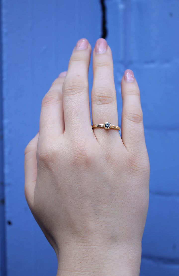 A left hand with light skin tone wears the Madeleine Azure Drop 9ct Yellow Gold Ring, featuring a small round stone on the ring finger. The short fingernails are polished light pink, set against a blue background.