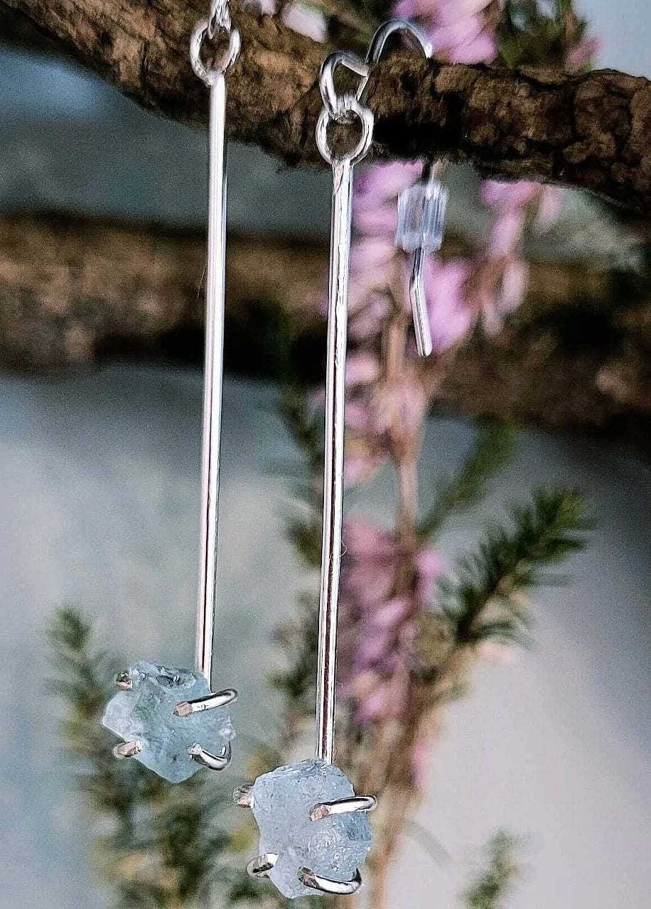 Close-up of a pair of Raw Aquamarine Drop Earrings by Caroline Stokesberry-lee with sterling silver bars and raw aquamarine gemstones at the ends. The earrings hang from ear wires on a branch with blurred green foliage and pink flowers in the background.