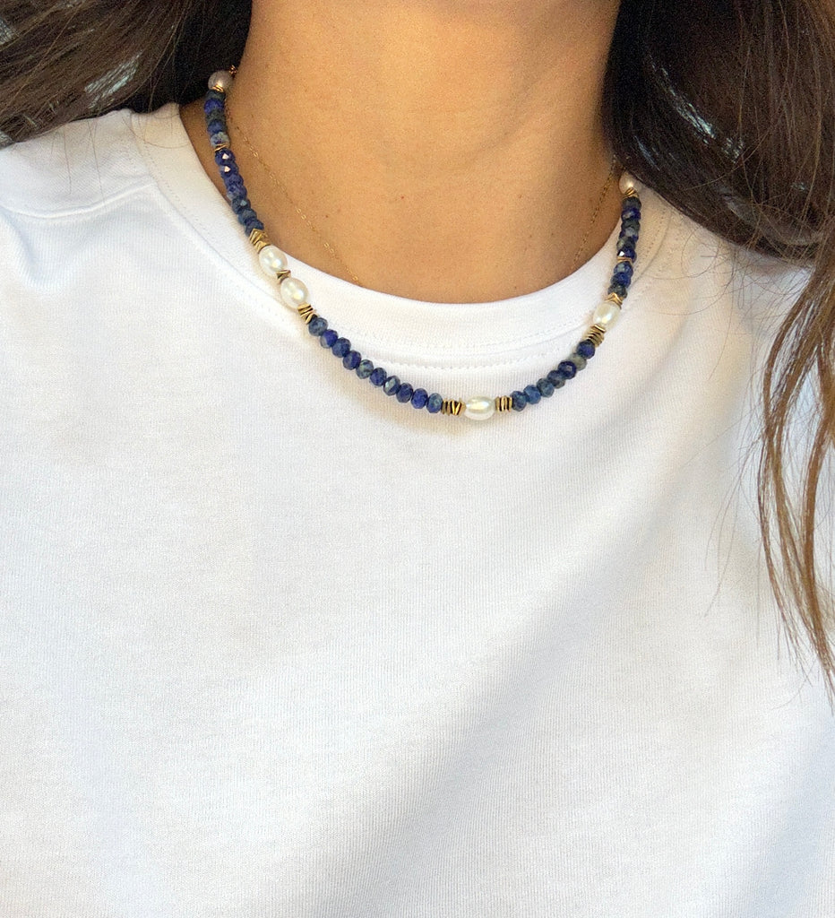 A person wearing a white crew-neck shirt and the Aria V Ariel Necklace, a handmade choker with dark blue lapis lazuli, gold, and white beads. The photo is cropped to highlight the necklace and evokes subtle Dublin jewelry style.