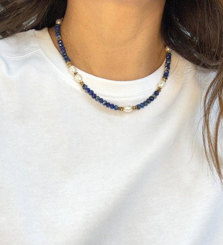 A person wearing a white crew-neck shirt and the Aria V Ariel Necklace, a handmade choker with dark blue lapis lazuli, gold, and white beads. The photo is cropped to highlight the necklace and evokes subtle Dublin jewelry style.