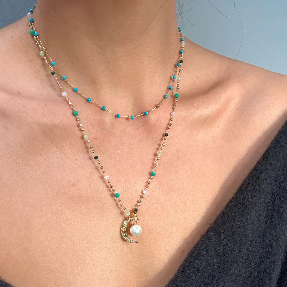 A person wears two handmade necklaces by Aria V: a short strand of blue beads and the Aurelia Necklace—a longer moon gold chain with a pearl—both crafted in Dublin. They pair them with a dark off-shoulder top.