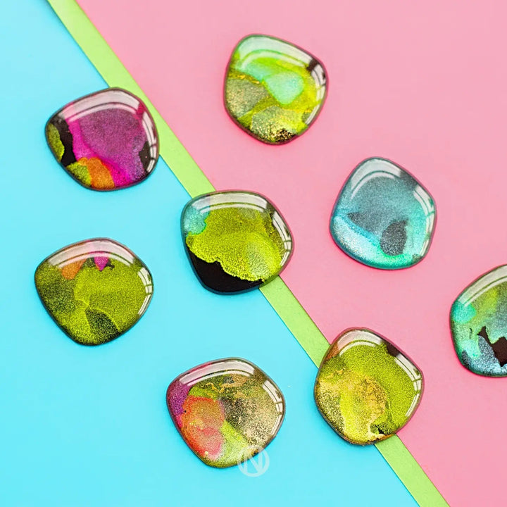 A collection of colorful, unique resin earrings arranged on a split background of pastel blue and pink with a thin green strip in between. These abstractly painted pieces feature hues of green, gold, pink, and blue are the Aurora Stud Earrings by Naoi.