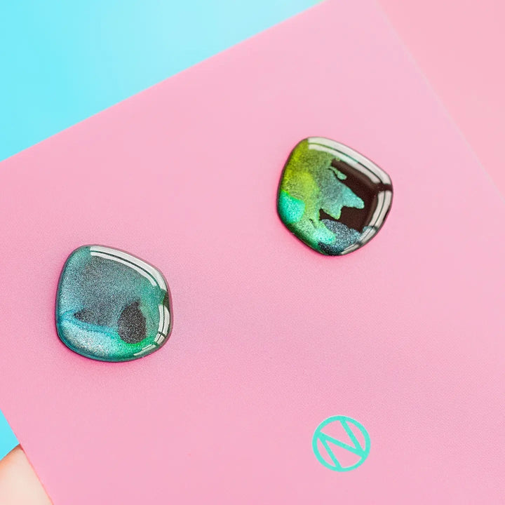 A pair of iridescent, asymmetrical, hand-painted Aurora Stud Earrings by Naoi in shades of green, blue, and black are displayed on a pink card with a green logo in the bottom center. The background is a gradient of light blue to pink.