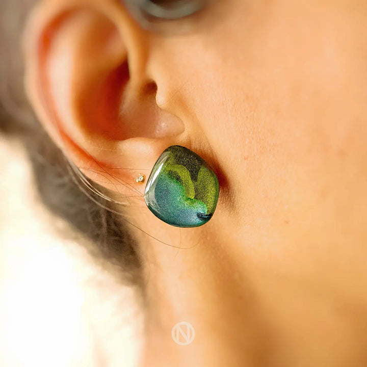 Close-up of a person wearing Naoi's Aurora Stud Earrings with iridescent green and blue hues on their left ear. The shiny, cubic-like shape stands out against the person's skin, anchored by stainless steel earring posts. The background is softly blurred, focusing on the striking earrings.