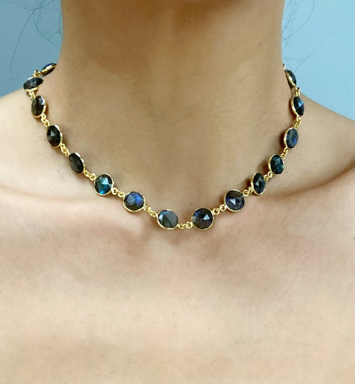 The Aurora Necklace by Aria V is a delicate piece, featuring round, dark-colored labradorite stones set in gold bezels. Each stone has a slight iridescent quality, giving off subtle hints of blue and green when hit by light. This handmade jewelry is worn around the neck and photographed against a plain, light blue background.