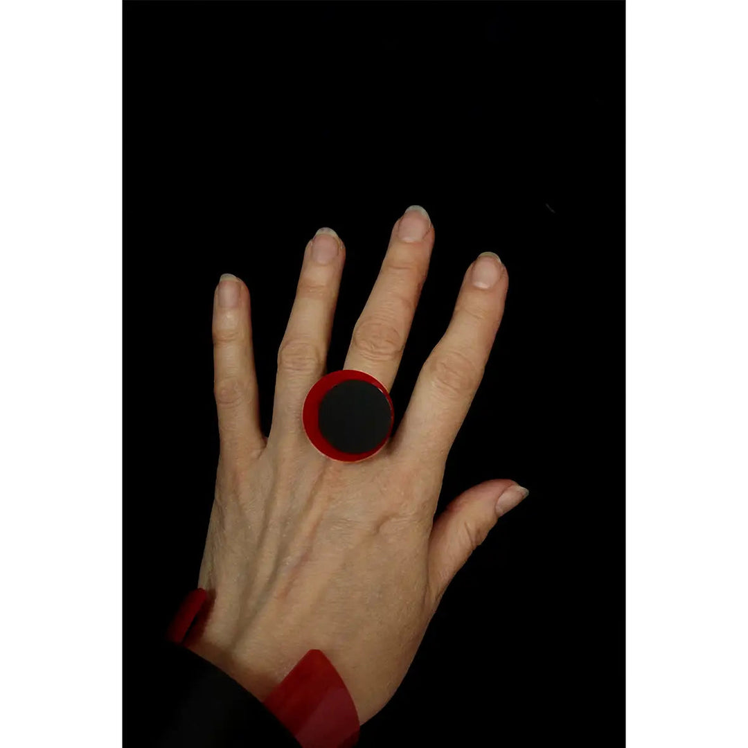 A hand wearing a large, round, red and black Amy Ring by Alex + Svet on the middle finger is shown against a black background. The fingers are slightly spread, displaying the handcrafted jewelry prominently.