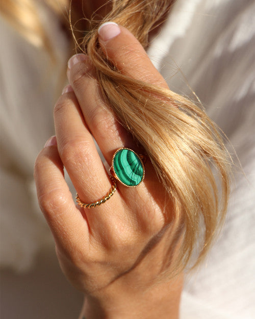 A close-up of a hand holding blonde hair strands. The hand is adorned with a twisted gold ring and a larger Nilai Frida Ring featuring a green natural stone with gold detailing. The person is wearing a white, textured garment in the background.