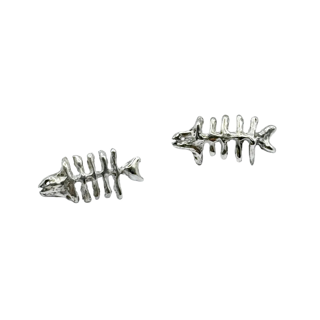 Two small, metallic Baraka Earrings by Soufana Attia, shaped like fish skeletons with a head, spine, ribs, and tail, rest on a plain light gray background. Their design echoes Arab symbols found in Baraka jewelry and protection traditions.