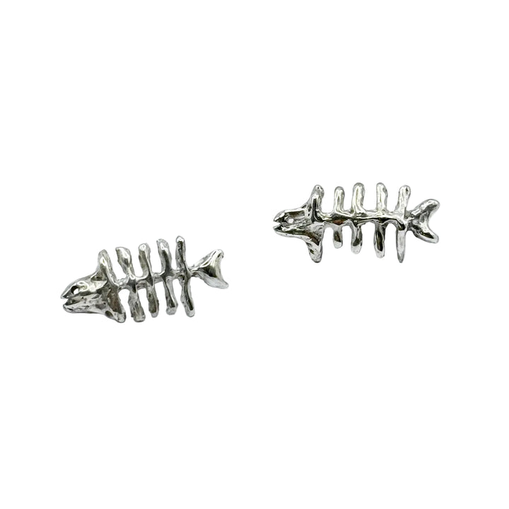 Two small, metallic Baraka Earrings by Soufana Attia, shaped like fish skeletons with a head, spine, ribs, and tail, rest on a plain light gray background. Their design echoes Arab symbols found in Baraka jewelry and protection traditions.