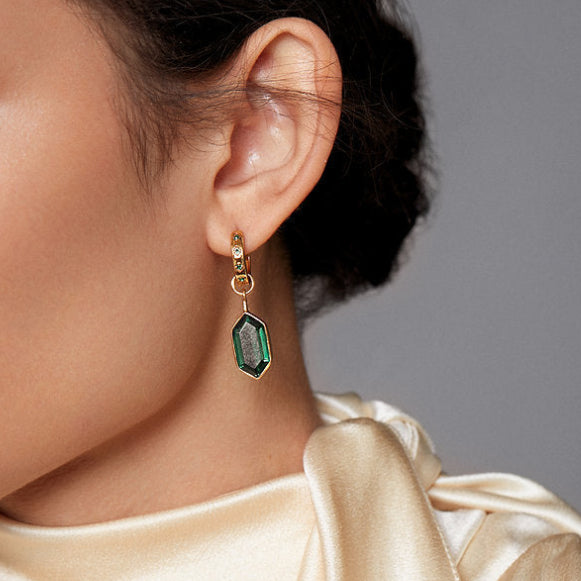 A close-up of a model's ear wearing a gold hoop earring with a dangling green faceted gemstone charm.