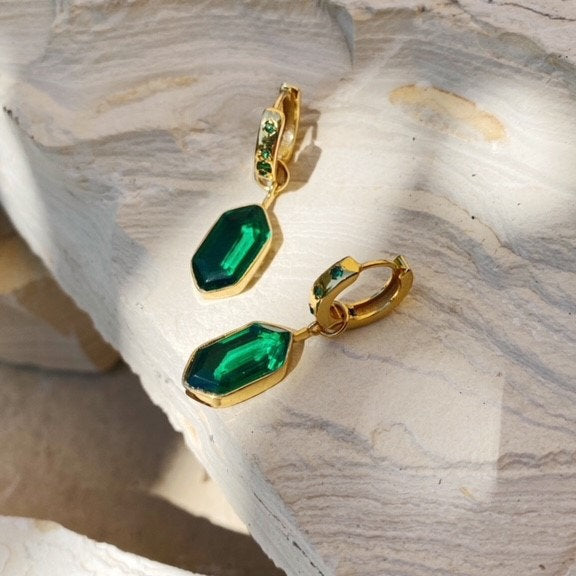 Gold hoop earrings with large faceted green gemstone drops, displayed on a textured white stone surface in bright natural light.