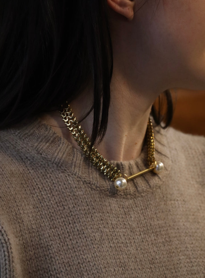 A person wears the Zubeldia Gold Bar and Double Pearl choker, featuring two large pearls, paired with a beige knit sweater. Photographed side-on from the neck up, their dark hair is partially visible.