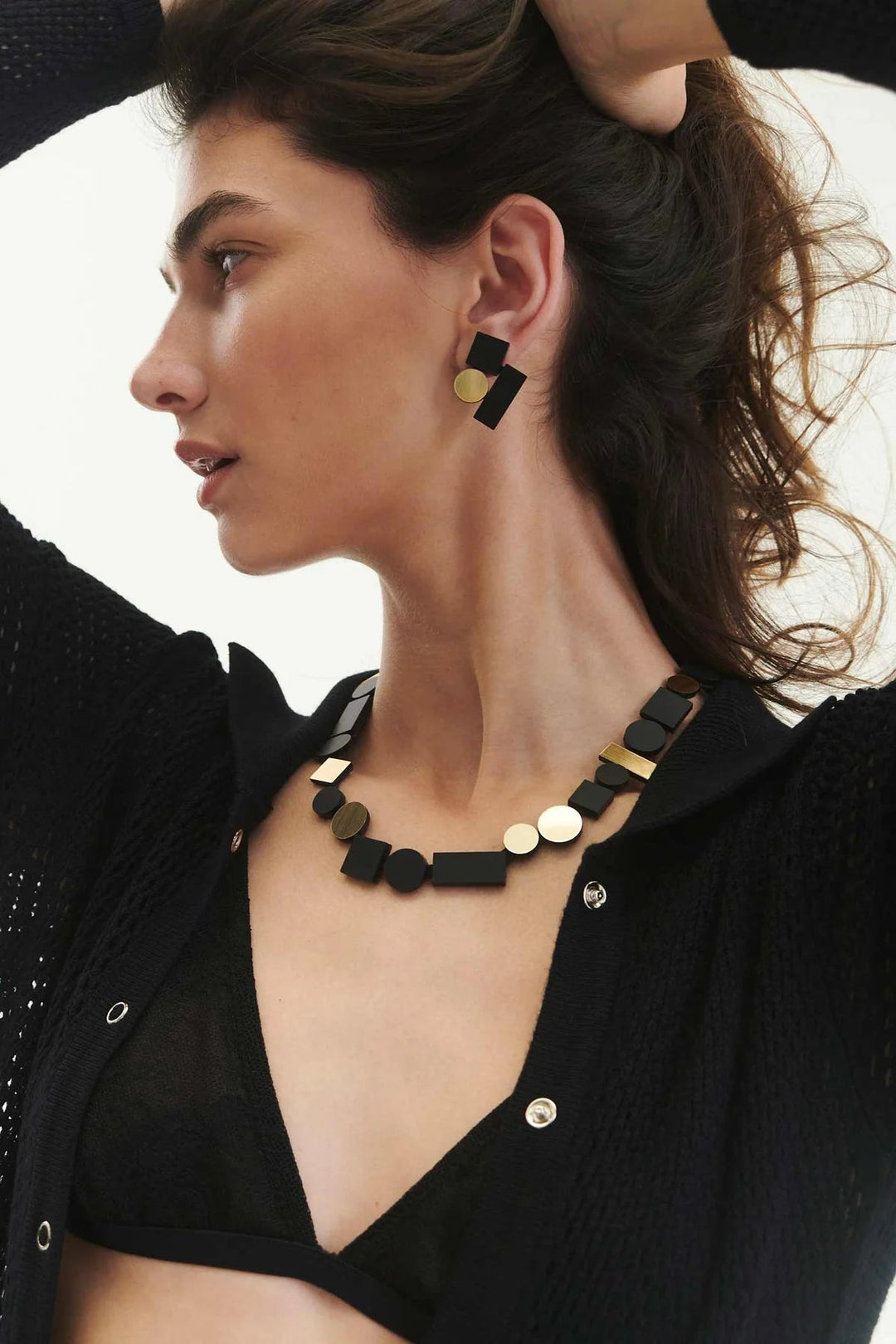 A woman with wavy brown hair wears geometric black and gold Bauhaus Earrings Small from the Iskin Sisters collection and a matching necklace. She is dressed in a black textured shirt over a black bralette, with her left hand raised to her head and her face in profile, looking to the left.