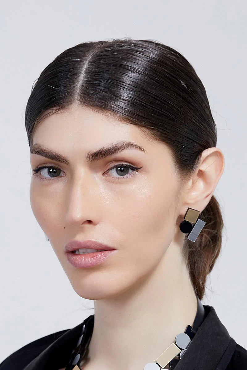 A woman with dark, slicked-back hair and light skin wears the Iskin Sisters' Bauhaus Earrings Small in black, known for their material excellence and geometric metallic design. She has a serious expression and is dressed in a black outfit against a plain, light-colored background.