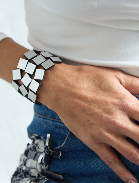 A close-up of a person's wrist wearing a chunky geometric bracelet made of alternating silver squares, styled with a white t-shirt and blue jeans.