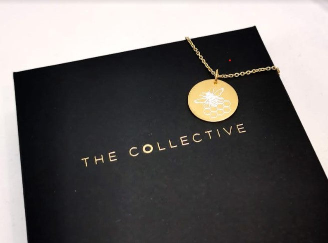 A gold-plated necklace with a round pendant featuring a bee design rests on a black box with the gold-embossed text "The Collective Dublin." The box sits on a light surface.