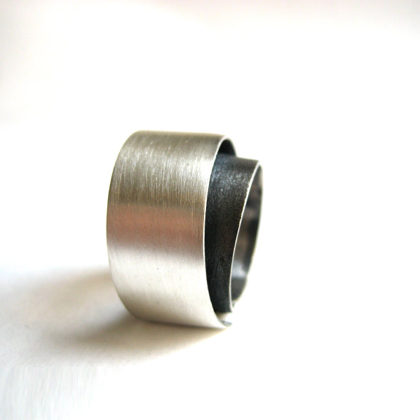 A silver, wide-band ring with a modern, minimalist design is shown. Part of the MarCucurella Bilbao Silver Ring collection, the ring features a layered, overlapping band with a brushed matt silver texture on the outer layer and a smooth, darker-toned inner layer. The white background makes the ring the focal point.