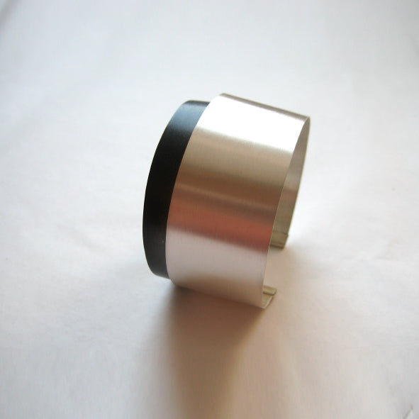 A modern, minimalist cuff bracelet from the Bilbao collection, featuring a smooth oxidized silver finish and a black accent on one side. Displayed on a white surface, this handmade jewelry piece boasts a sleek, open design and a slightly curved shape. The *Bilbao Bracelet* by *MarCucurella* exemplifies elegance and contemporary style.