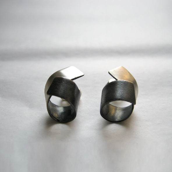 A pair of artistic, dark-toned metal rings with abstract, angular designs from the Bilbao collection. Each handmade Bilbao Silver Earrings by MarCucurella features unique, overlapping geometric shapes on top, showcasing a modern, sculptural style against a simple, light-colored surface.