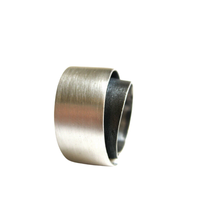 A close-up image of a cylindrical metal ring with a brushed, matt silver finish on the exterior. From the MarCucurella Bilbao Silver Ring collection, the ring has an overlapping seam, giving it a slightly conical shape. The interior of the ring is dark and oxidized silver, contrasting with the shiny outer surface.