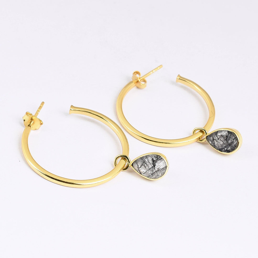 A pair of Black Routile Open Hoop Earrings by Watermelon tropical with dangling, teardrop-shaped charms that feature a marbled black and white pattern reminiscent of Black Routile stone. The earrings are laid flat on a white surface.