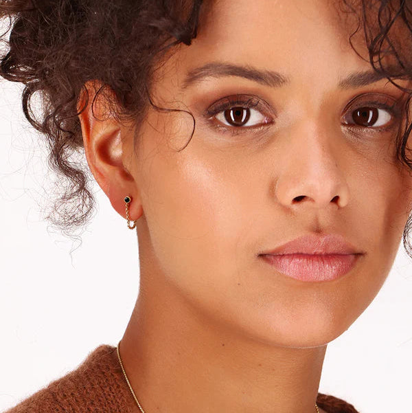 A woman with curly brown hair and glowing skin looks at the camera, wearing Scribble & Stone's Black Chain Stud Earrings and a brown top. The background is plain white.
