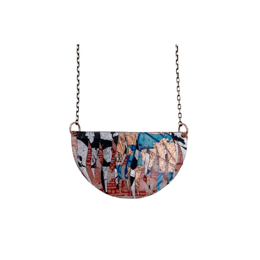 A vibrant Bláth Batik Textile Necklace by Rothlú, featuring a semi-circular pendant, hangs from a sleek dark chain. This handmade piece showcases an abstract design with hues of orange, copper, silver, blue, and black, reminiscent of a collage of torn paper or paint splatters. Its unique texture is elegantly accentuated by an eco resin finish.