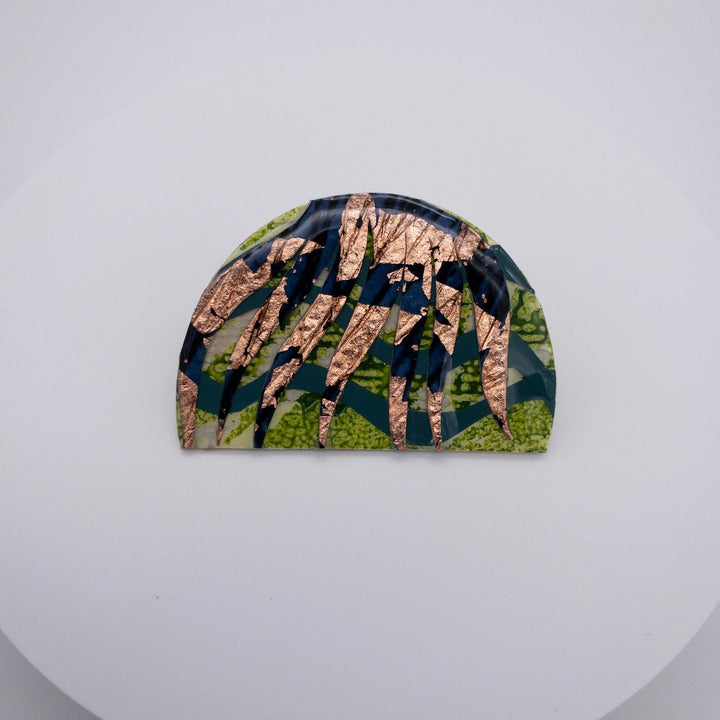 A semi-circular eco resin brooch with an intricate design featuring green, black, and gold patterns. The hand-patterned Bláth Batik Textile Brooch by Rothlú showcases irregularly shaped fragments forming an abstract mosaic-like appearance. The unique textured brooch is displayed on a plain white surface.