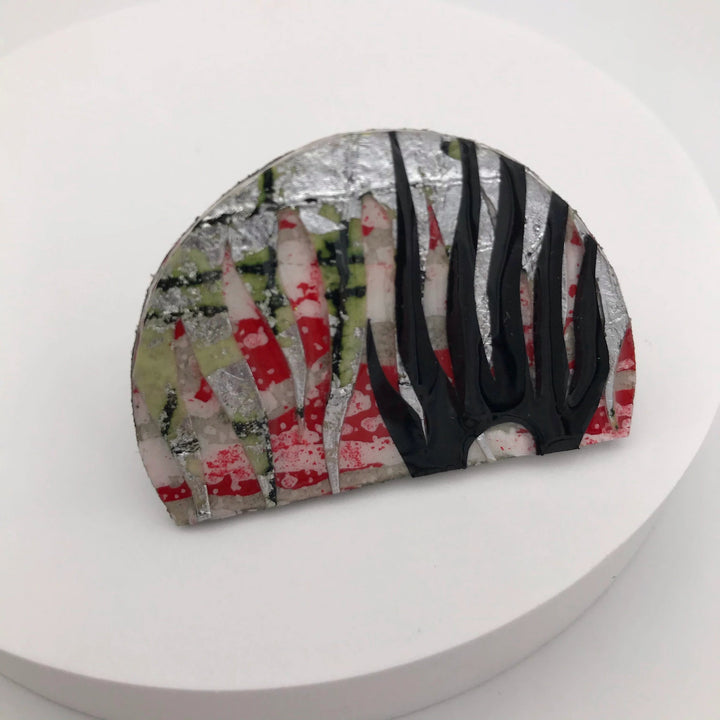 A unique Bláth Batik Textile Brooch by Rothlú shaped in a semi-circle, featuring a mix of red and white stripes, green hues, and a silver foil-like texture. Black vertical lines resembling flames or tall grasses extend from the right side, all on a pristine white background.