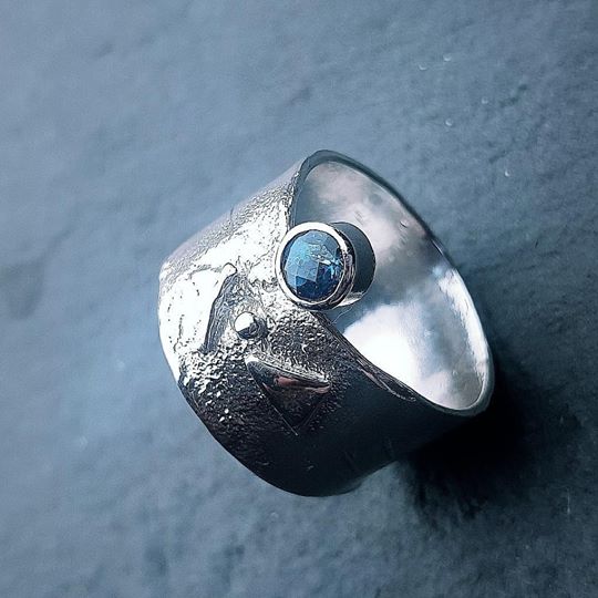 A wide sterling silver ring featuring a textured surface with an embedded round Blue Topaz gemstone and abstract engraved designs on the band. The background is a soft-focus, dark grey surface, highlighting the elegance of this Blue Topaz Guardian Ring by Caroline Stokesberry-lee.