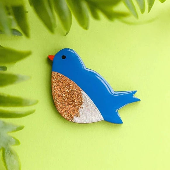 A small, handpainted wooden Bluebird Pin Brooch by Naoi with a glittery gold and beige belly is pinned against a bright green background. Green leaves frame the corners, adding a natural touch to the scene.