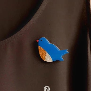 A Naoi Bluebird Pin Brooch with an orange beak and a wing featuring gold and white glitter is pinned to black fabric, all elegantly secured by a brass butterfly pin.