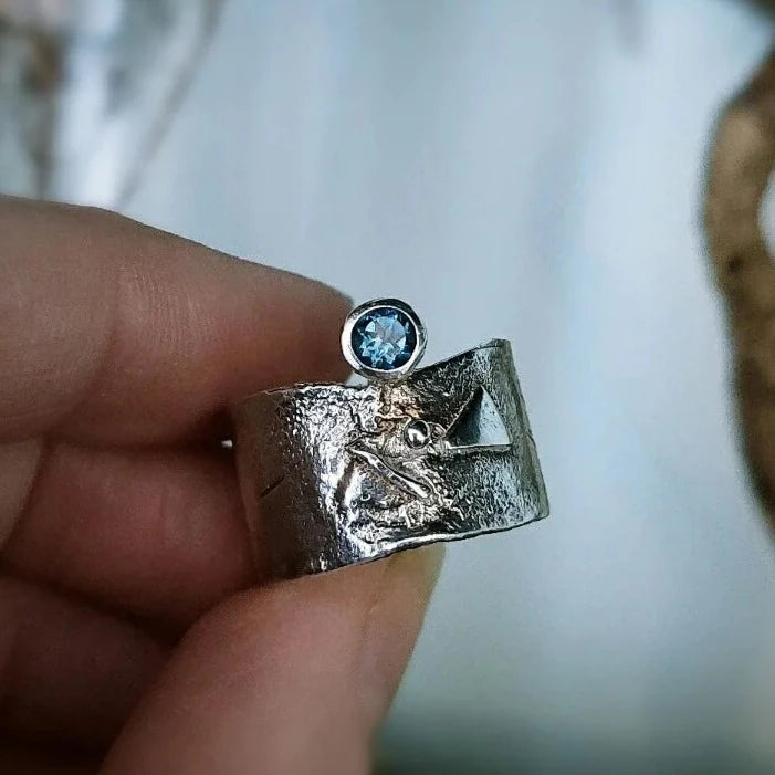 A close-up of a hand holding a Caroline Stokesberry-lee Blue Topaz Guardian Ring featuring a textured design with a small, round Blue Topaz gemstone set on top. The ring has a unique, sculptural appearance.