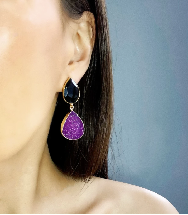 Close-up of a woman's ear adorned with the elegant Black Magic Earrings by Aria V, featuring a black teardrop-shaped stone at the top and a larger purple Druze stone outlined in 16k gold plated brass at the bottom. The background is softly blurred in gradient gray.