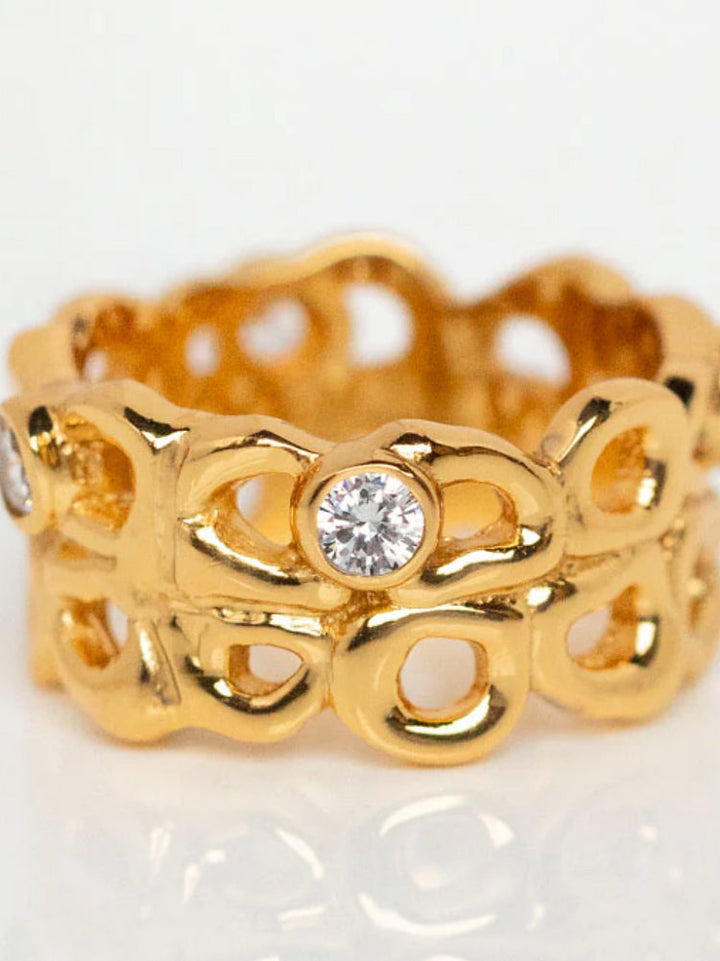 An intricately designed handmade gold ring featuring an openwork pattern with rounded shapes. The design includes several small, round, clear gemstones set throughout the ring, adding a touch of sparkle and elegance. This exquisite piece is known as the Bordeaux Ring from Shyla. The background is plain white.