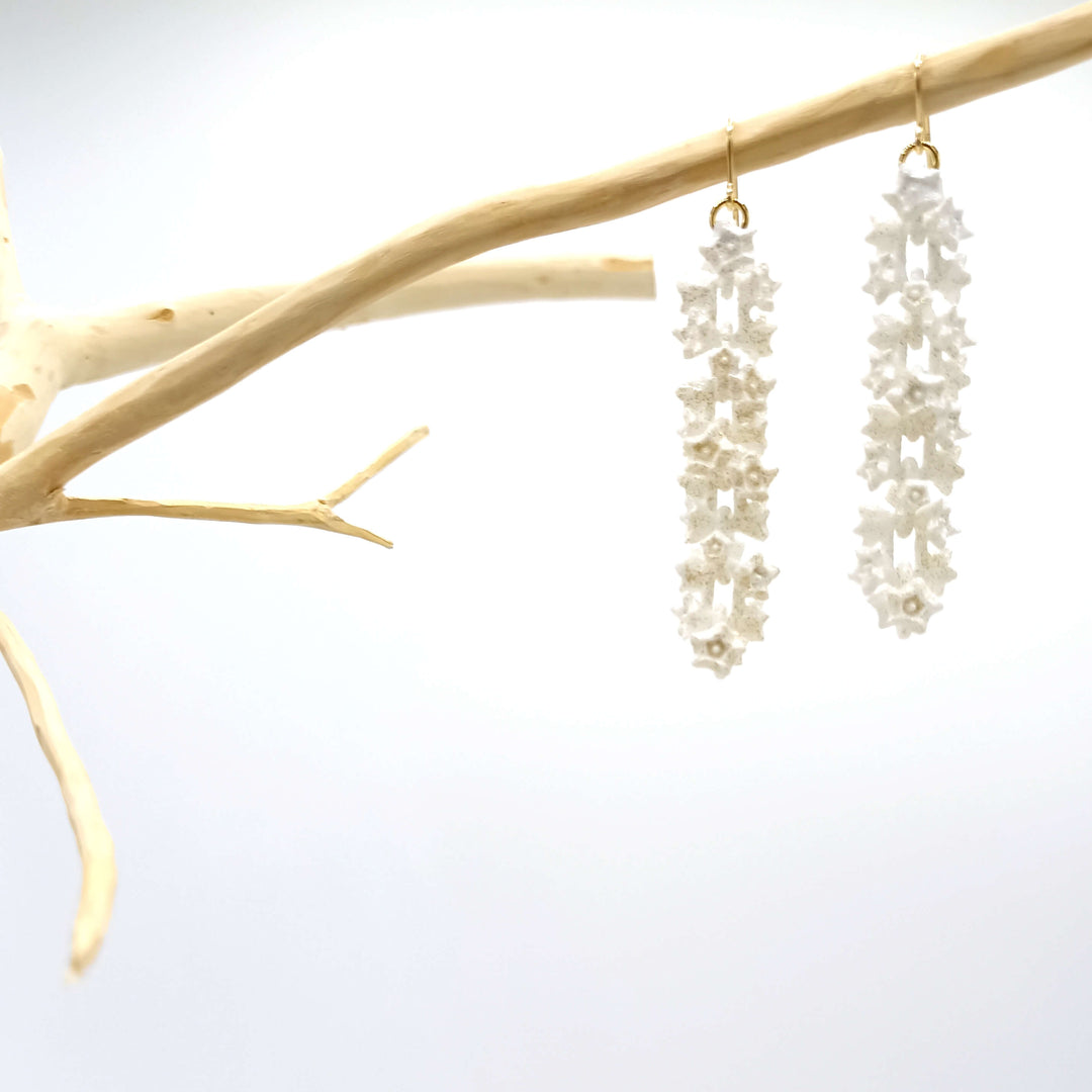 A pair of intricate, White Sakura Earrings by Tochu-De hang from a beige, minimalist branch against a white background. The 3D printed jewellery design features connected ornate floral patterns in gold stainless steel, creating a delicate and elegant appearance.