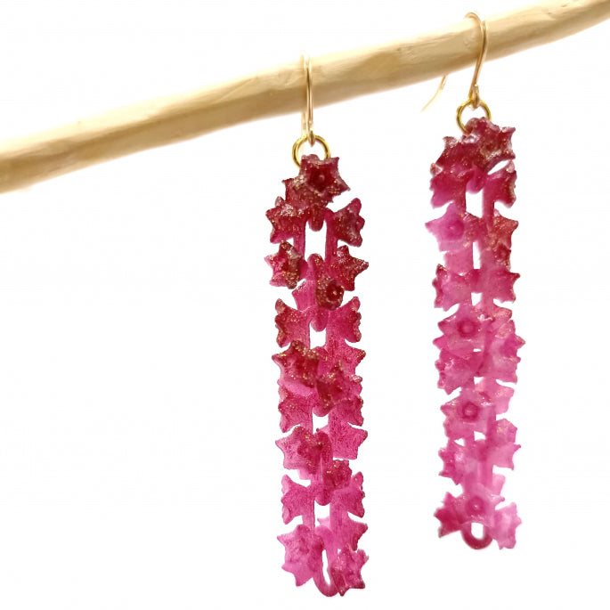 A pair of long, dangling Fuchsia Sakura Earrings made of pink, flower-shaped beads hanging from a bar. The earrings are vibrant and intricate, with a shimmering finish—perfect floral accessories for any occasion.