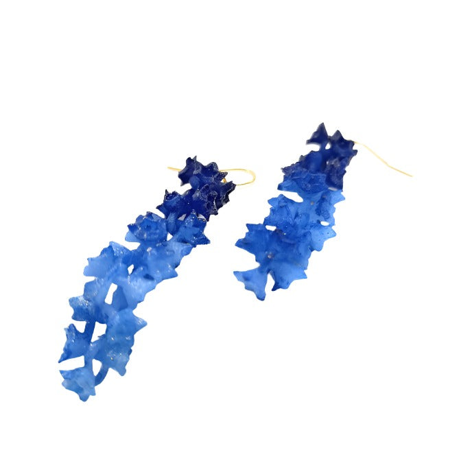 A pair of long earrings featuring a cascading design of blue and deep blue crystal-like material. The Blue Sakura Earrings, part of the Tochu-De SAKURA Flower Jewelry collection, are arranged on a white surface, emphasizing the varying shades of blue and the intricate, star-shaped crystalline structure.